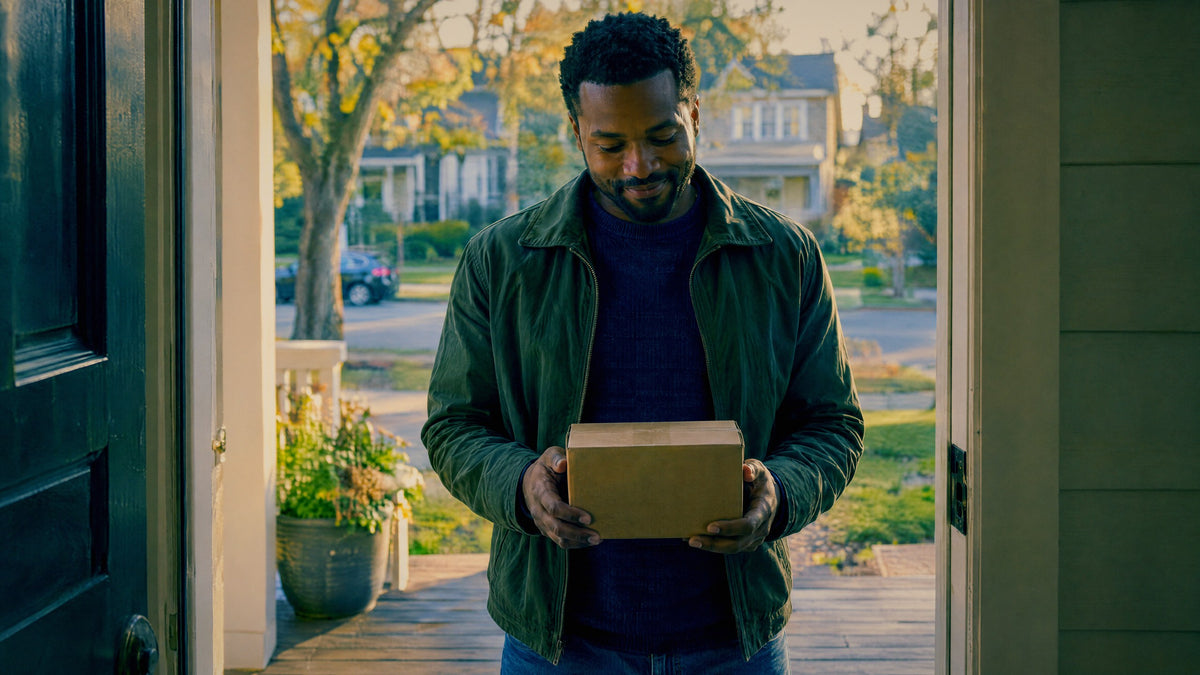 A man stands in his open doorway holding a small plain parcel he has just collected from the step.