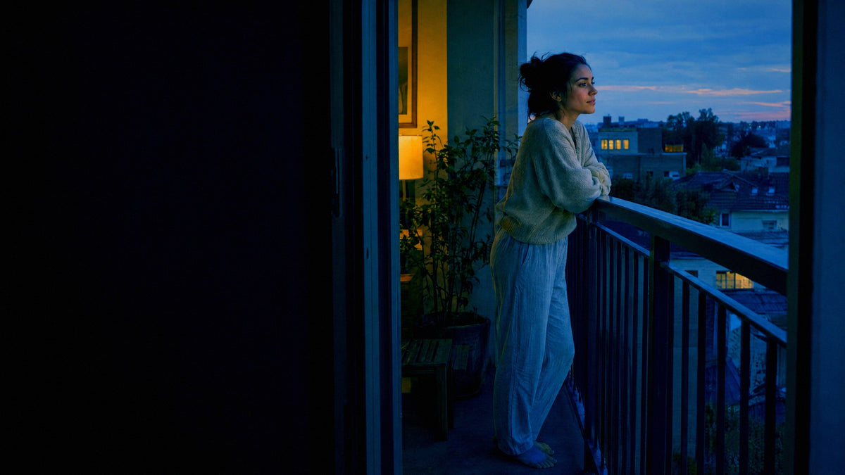 A woman stands at the open door of her apartment balcony at dusk, looking out over the neighbourhood