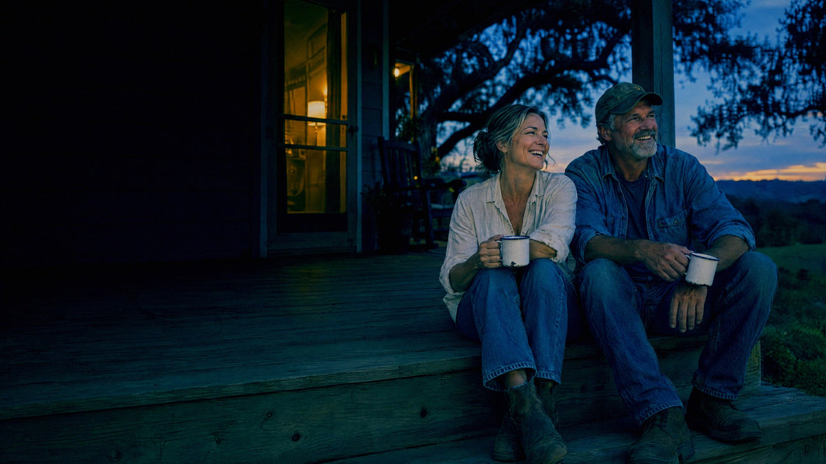 A couple sit laughing on the steps of a farmhouse porch at blue hour with mugs in hand