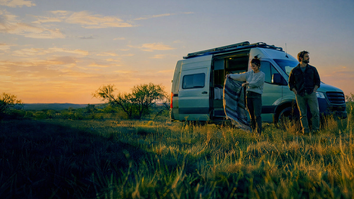 A couple shake out a blanket beside a camper van on open ranch land at golden hour