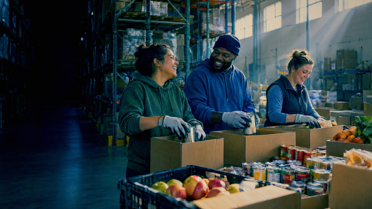Three volunteers pack boxes at a long table in a community food pantry, daylight cutting across the space