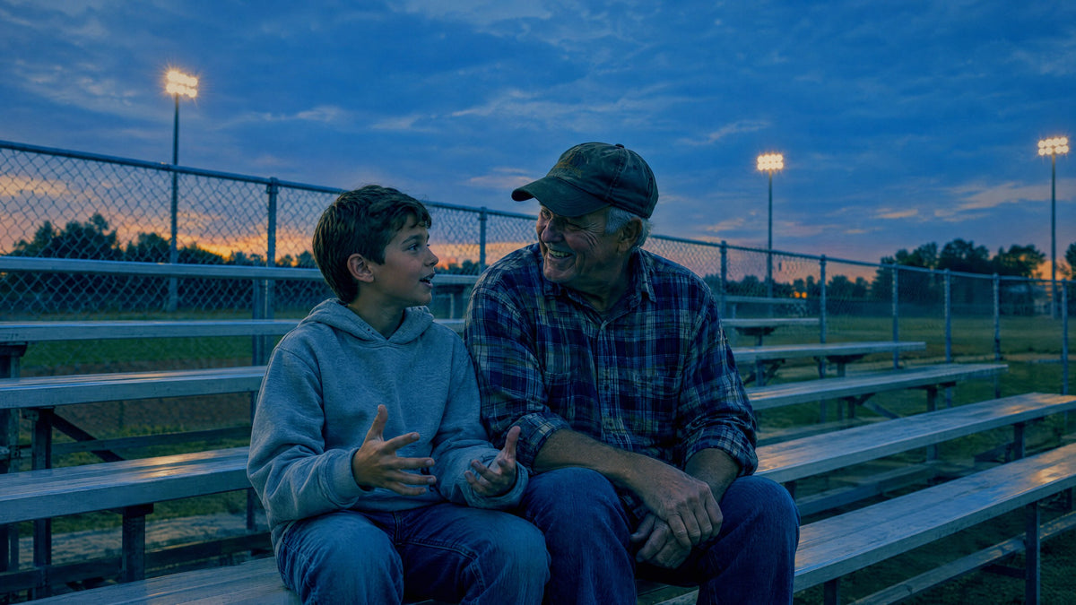 A grandfather laughs with his grandson on the bleachers of a small town ball field at dusk.