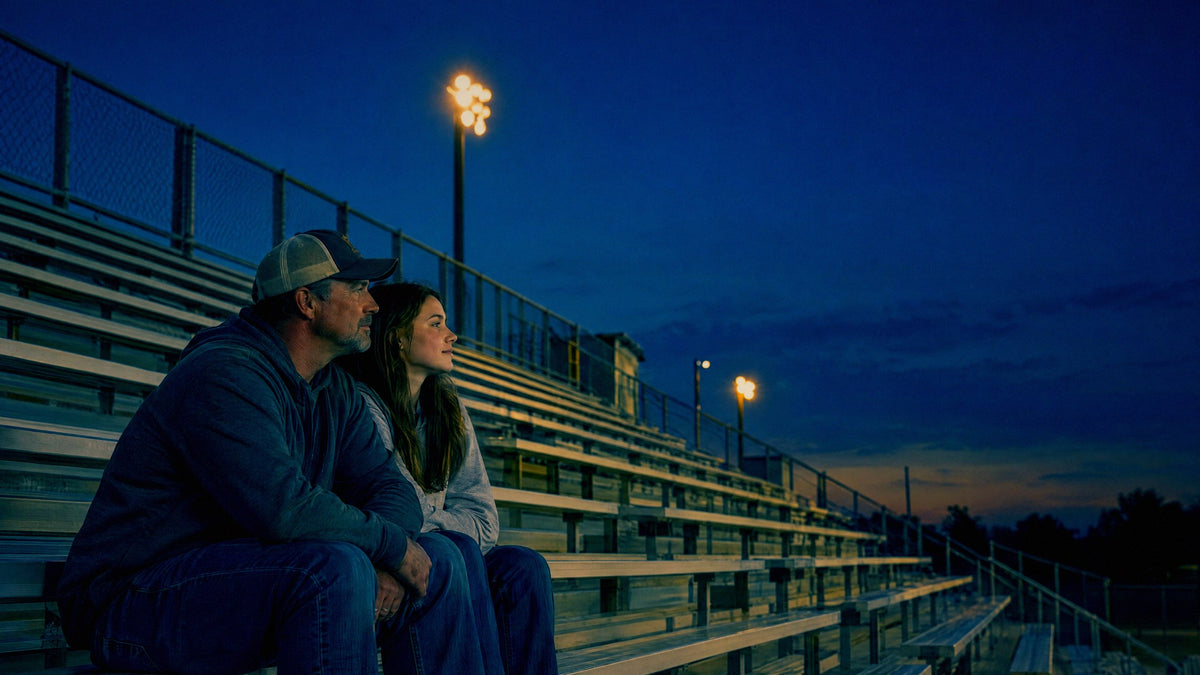 A father and daughter sit together on empty bleachers at a small town ball field at dusk