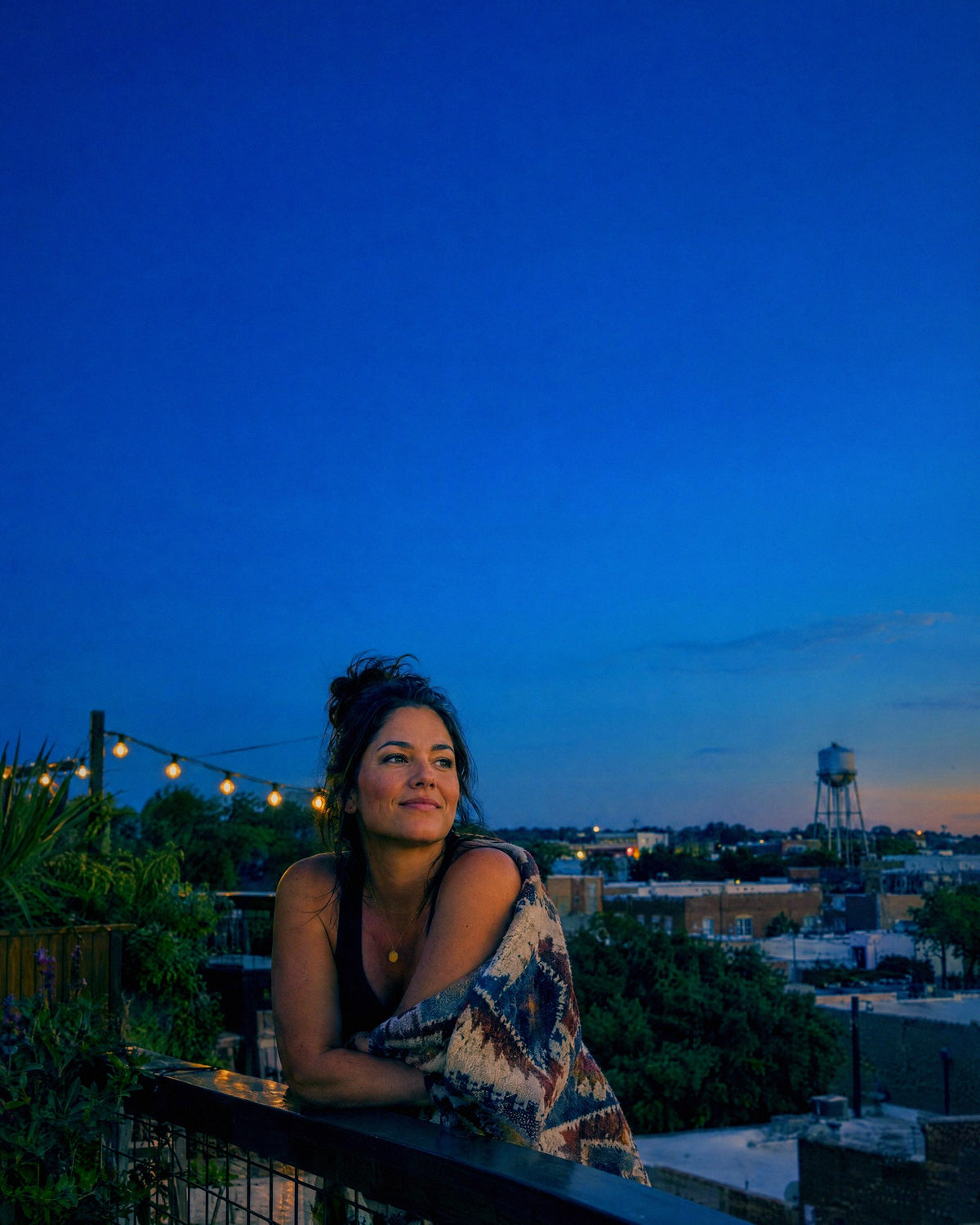 A woman leans on a rooftop garden railing in the early evening above a low brick neighbourhood